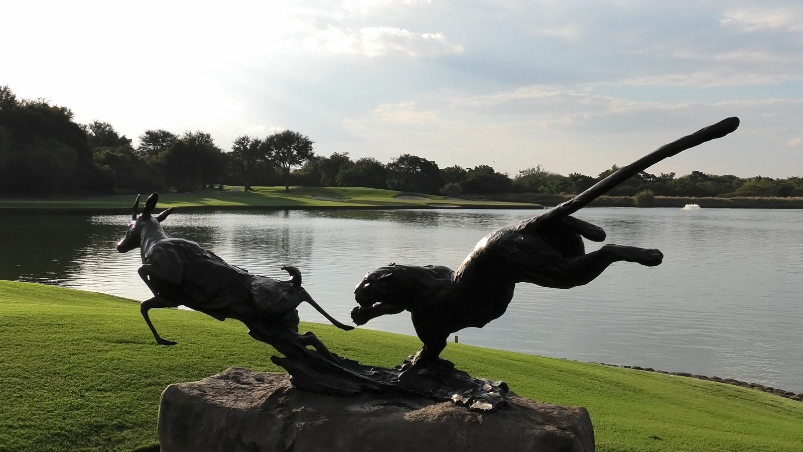 Bronze leopard and antelope sculpture beside a golf course lake at dusk